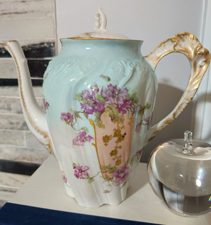 La tetera de Chamart Limoges France mide 18cm de alto y está elaborada en porcelana pintada a mano, destacando un elegante motivo floral en tonos rosa y lavanda, con detalles dorados sobre un fondo verde menta pálido.