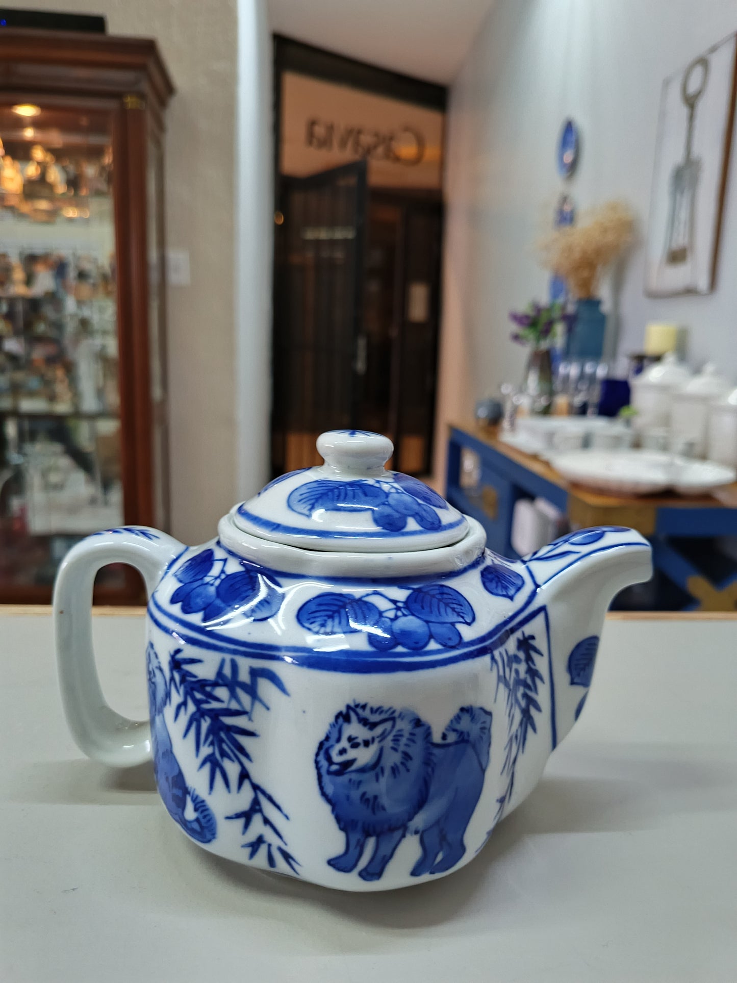 Tetera vintage de porcelana azul y blanca presenta un diseño elegante de perros y árboles frutales, complementada por una forma de flor.
