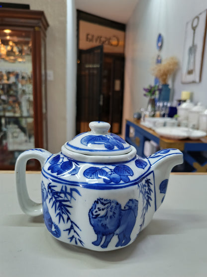 Tetera vintage de porcelana azul y blanca presenta un diseño elegante de perros y árboles frutales, complementada por una forma de flor.